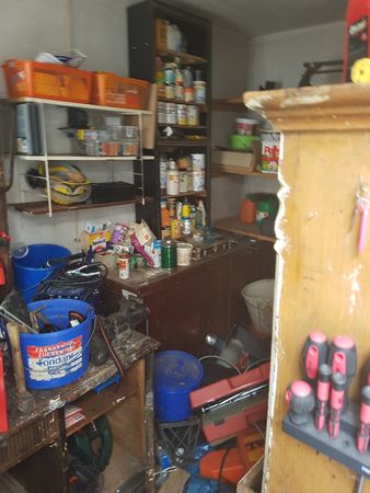 Unaufgeräumte Werkstatt mit Regalen, Werkzeug, Farbeimern und verschiedenen Utensilien.